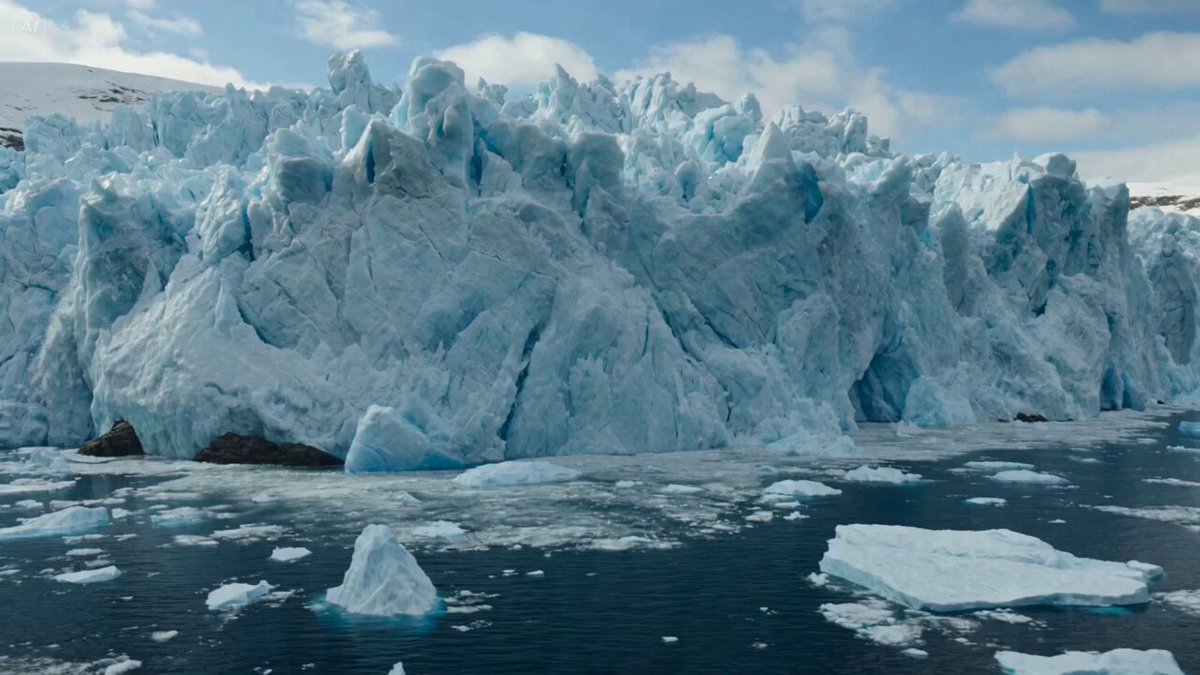 A giant glacier wall collapses into a fjord beside a coastal city.