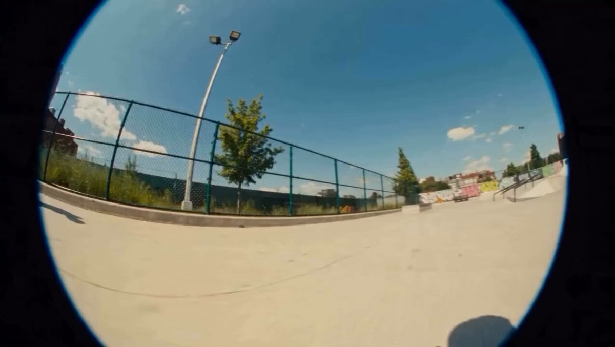 Fisheye lens skatepark shot on a sunny day with chain-link fences and graffiti-covered ramps in 